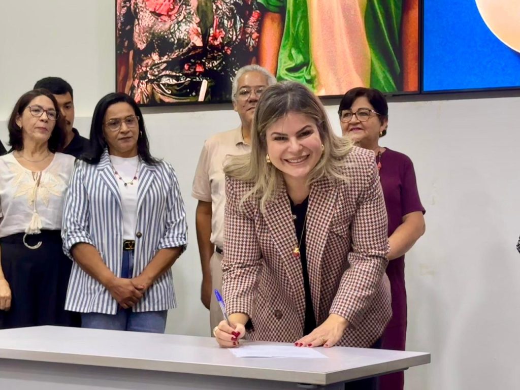 Close-up de uma mulher loira, sorridente, vestindo um blazer xadrez em tons de marrom e bege. Ela está inclinada sobre uma mesa branca, assinando um documento com uma caneta azul. Ao fundo, em pé, observa-se um grupo de pessoas de forma desfocada, incluindo homens e mulheres que acompanham o ato solene.