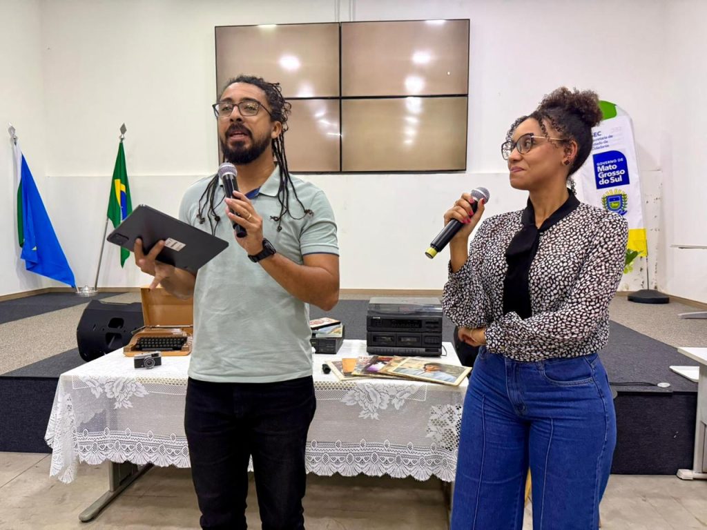 Um homem e uma mulher em pé no palco atuando como apresentadores. O homem, à esquerda, tem cabelos com tranças longas, usa óculos e segura um tablet e um microfone. A mulher, à direita, tem cabelos cacheados presos em um coque alto, usa óculos e também segura um microfone. Entre eles, uma mesa decorada com objetos retrô, como uma máquina de escrever, rádio antigo e discos de vinil.