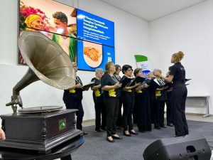 Fotografia em plano médio de um coral formado por mulheres idosas. Elas vestem camisetas pretas com um detalhe amarelo na lateral e seguram pastas de partitura pretas. À direita, uma jovem regente de costas, vestindo roupa preta e com cabelos cacheados presos, conduz o grupo. No canto inferior esquerdo, destaca-se um gramofone antigo sobre uma mesa. Ao fundo, um painel digital exibe a identidade do "1º Fórum Conectando Gerações".