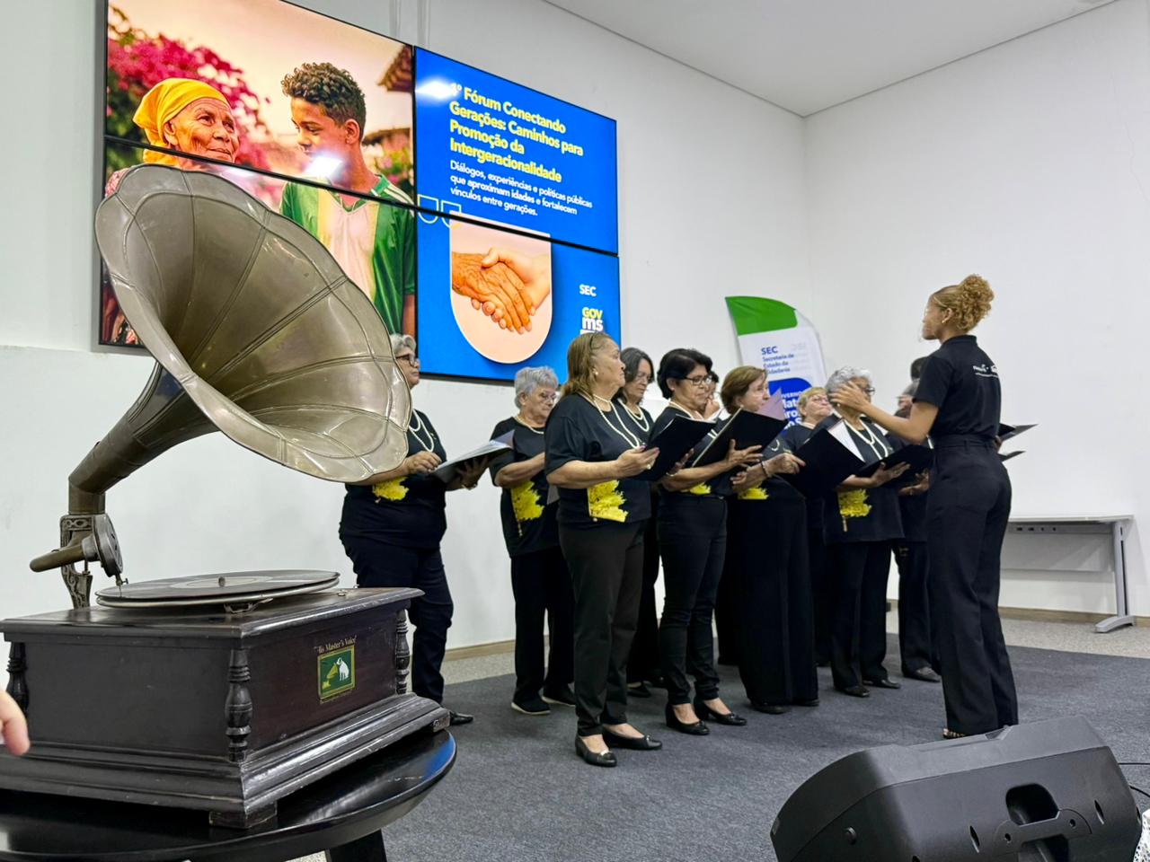 Fotografia em plano médio de um coral formado por mulheres idosas. Elas vestem camisetas pretas com um detalhe amarelo na lateral e seguram pastas de partitura pretas. À direita, uma jovem regente de costas, vestindo roupa preta e com cabelos cacheados presos, conduz o grupo. No canto inferior esquerdo, destaca-se um gramofone antigo sobre uma mesa. Ao fundo, um painel digital exibe a identidade do "1º Fórum Conectando Gerações".