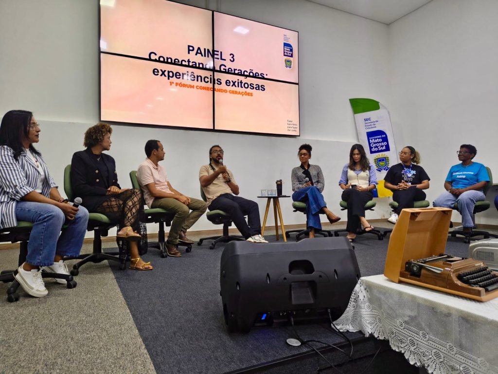 Registro de um painel de discussão com oito participantes sentados em semicírculo no palco. Entre os debatedores, há homens e mulheres de diferentes etnias e idades. No centro, um homem negro fala ao microfone. À frente deles, no chão, há uma caixa de som e uma mesa com uma máquina de escrever antiga sobre uma toalha de renda. O telão ao fundo anuncia o "Painel 3: Experiências Exitosas".