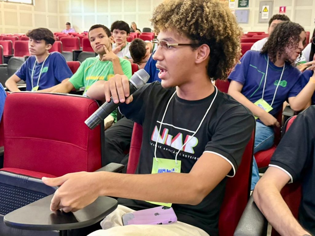 Um jovem de óculos e cabelos crespos descoloridos está sentado em uma poltrona vermelha de auditório. Ele segura um microfone com a mão direita e fala com expressão entusiasmada. Ele veste uma camiseta preta com a estampa "NIAR" e usa um crachá de identificação no pescoço. Ao fundo, outros jovens em poltronas vermelhas observam a fala.