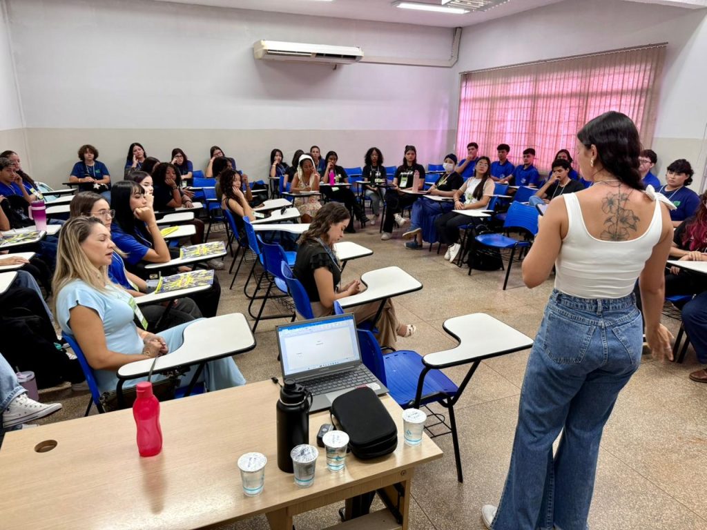 Uma mulher de regata branca e calça jeans, vista de costas e exibindo uma tatuagem floral, conduz uma atividade para um grupo de jovens em uma sala de aula. Os participantes estão organizados em semicírculo em cadeiras universitárias azuis. No primeiro plano à esquerda, uma mulher loira de blusa clara acompanha a dinâmica. Sobre uma mesa à frente, há um notebook aberto e garrafas de água.