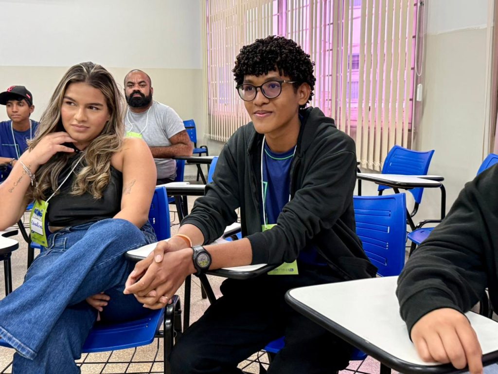 Em uma sala de aula ou de reuniões, um jovem negro com óculos e cabelos cacheados está sentado em uma cadeira azul com prancheta acoplada. Ele tem as mãos entrelaçadas sobre a mesa e olha atentamente para a frente. Ao seu lado, uma jovem com cabelos longos e luzes observa a mesma direção. No fundo, um homem barbudo e outros participantes completam a cena.