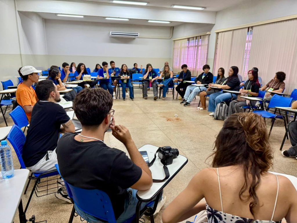 
O Gemini disse
Uma fotografia em plano médio mostra um grupo de cerca de 20 jovens reunidos em uma sala de aula, organizados em uma grande roda de conversa.