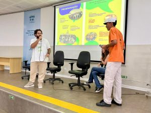 No palco de um auditório, dois jovens estão em pé diante de um telão que exibe a "Programação" do evento em tons de verde e amarelo. À esquerda, um homem de barba e óculos fala ao microfone. À direita, um jovem negro com boné e camiseta laranja segura um microfone e olha em direção a uma moça sentada em uma das cadeiras de escritório dispostas no palco. No fundo, há um painel da UFMS.