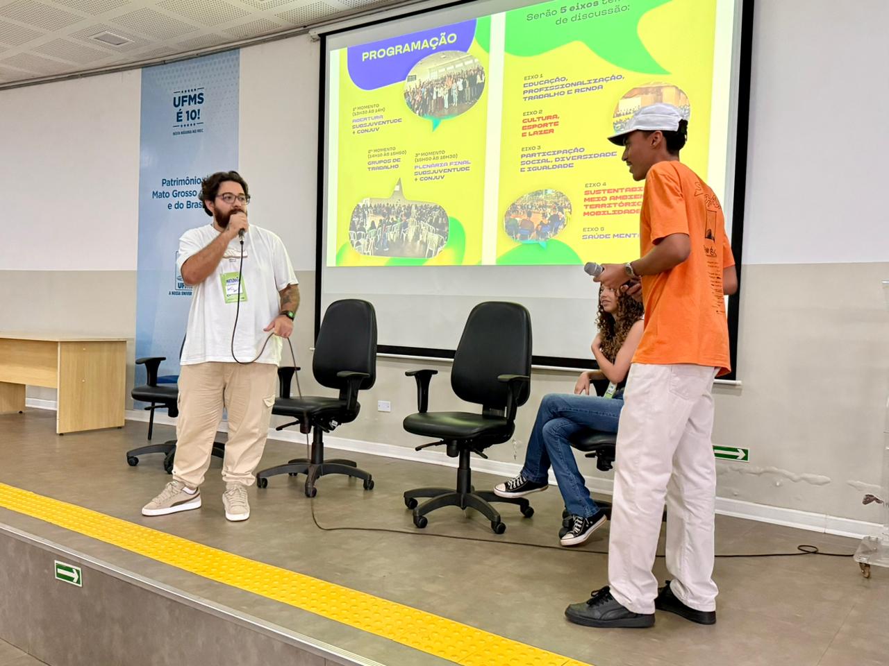 No palco de um auditório, dois jovens estão em pé diante de um telão que exibe a "Programação" do evento em tons de verde e amarelo. À esquerda, um homem de barba e óculos fala ao microfone. À direita, um jovem negro com boné e camiseta laranja segura um microfone e olha em direção a uma moça sentada em uma das cadeiras de escritório dispostas no palco. No fundo, há um painel da UFMS.