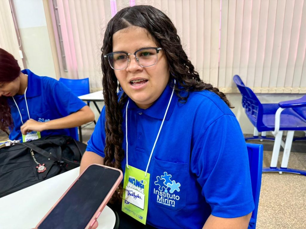 Plano fechado de uma jovem de óculos e longos cabelos ondulados. Ela veste a camiseta polo azul do Instituto Mirim e usa um crachá de identificação. Ela está com a boca levemente aberta, como se estivesse dando uma entrevista ou participando de uma discussão, enquanto um celular é segurado em frente a ela para gravar o áudio.