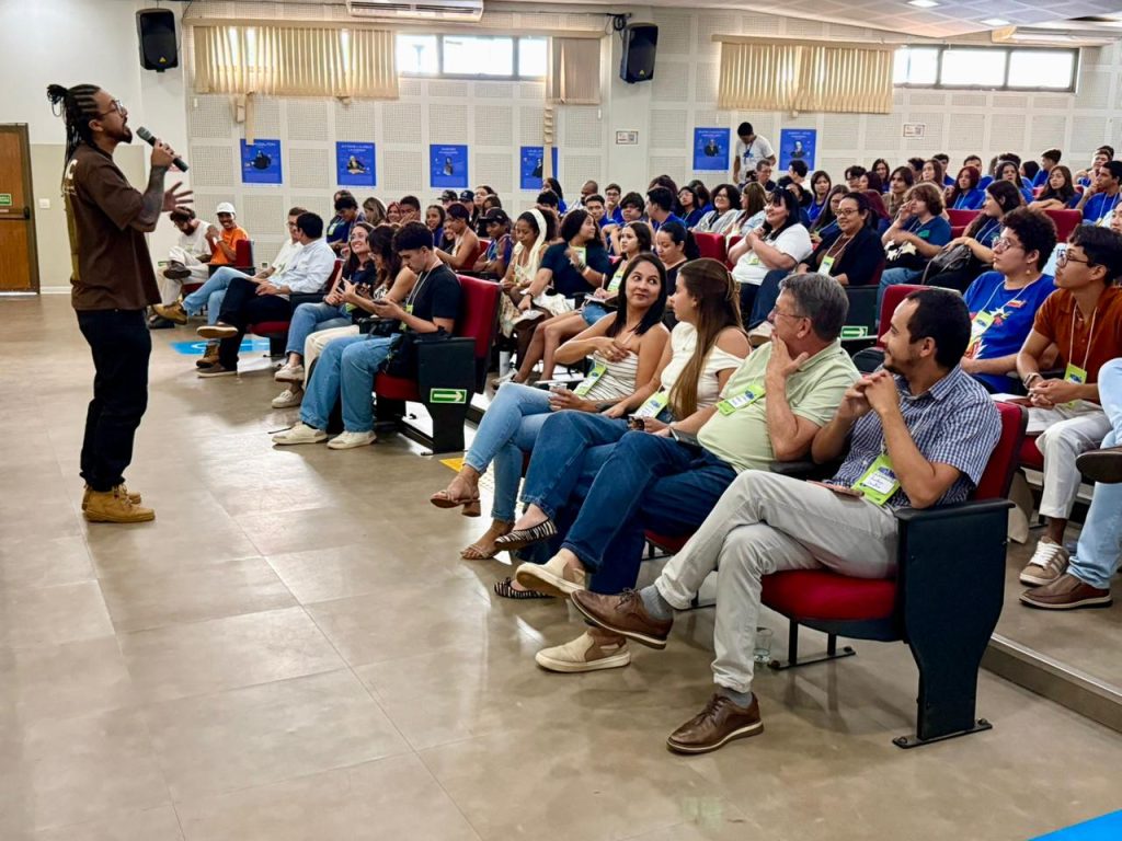Visão ampla do auditório lotado durante o Fórum das Juventudes. No lado esquerdo, um homem com dreadlocks e camiseta marrom fala ao microfone, de frente para o público. A plateia é composta por jovens e adultos sentados em poltronas vermelhas. O ambiente tem paredes claras com isolamento acústico e janelas altas com persianas. O clima é de atenção e participação.