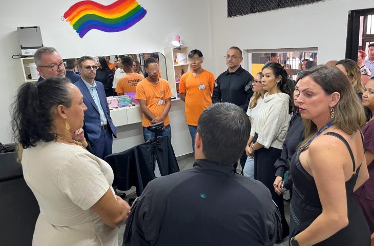 Autoridades, policiais e internos em uniformes laranjas reunidos em círculo dentro do salão de beleza durante a inauguração. O grupo conversa em um ambiente iluminado, próximo às bancadas de produtos.