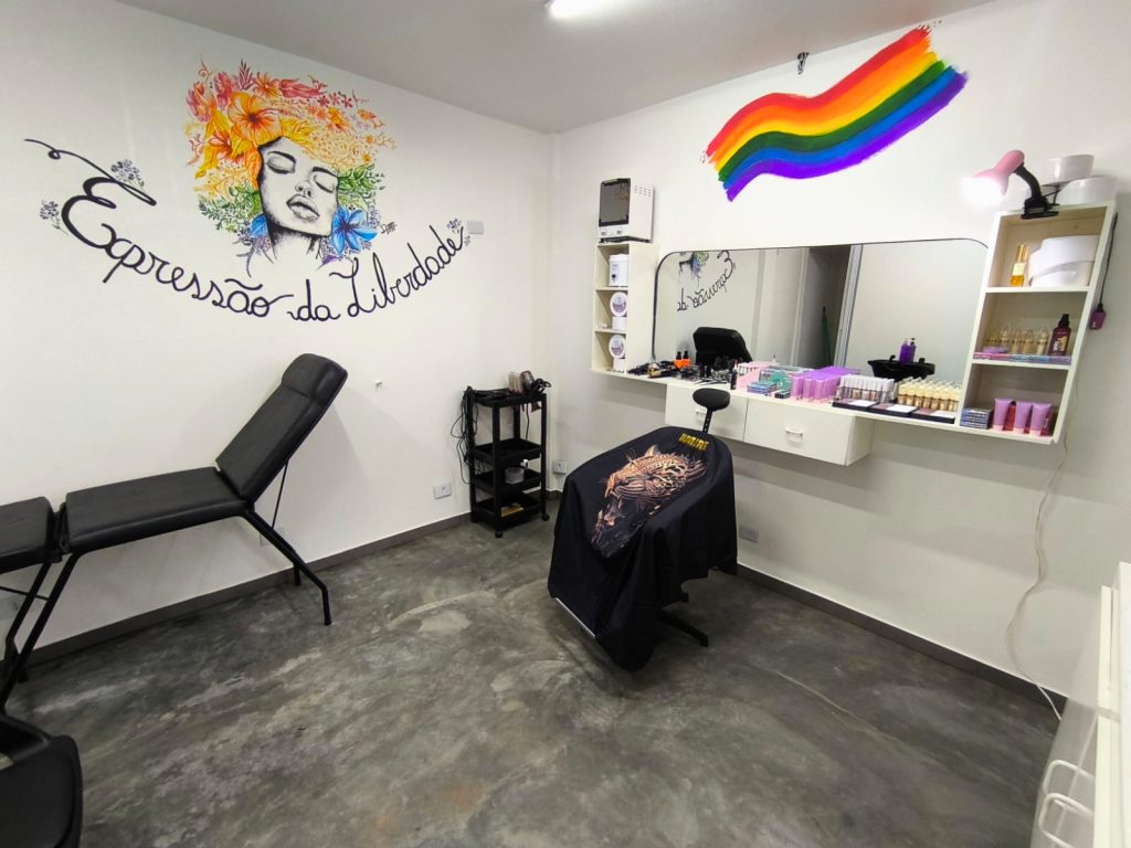 Interior de um salão de beleza com paredes brancas e piso de cimento queimado. Destacam-se uma maca preta, uma cadeira de cabeleireiro em frente ao espelho e pinturas na parede: o rosto de uma mulher com flores e uma bandeira do arco-íris.