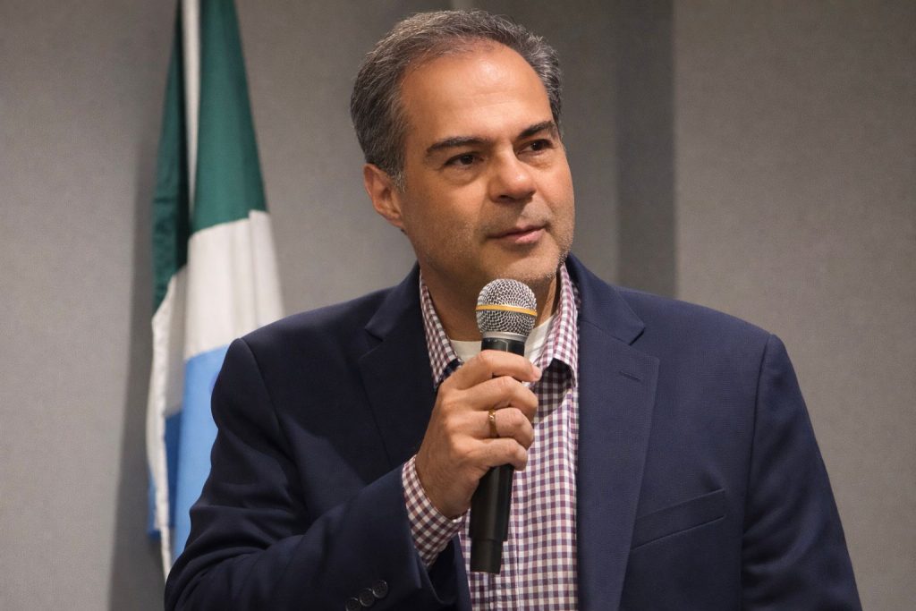 Homem branco de meia-idade, com cabelos grisalhos e paletó azul, fala ao microfone em um ambiente interno. Ao fundo, à esquerda, vê-se parte de uma bandeira com faixas branca, verde e azul.