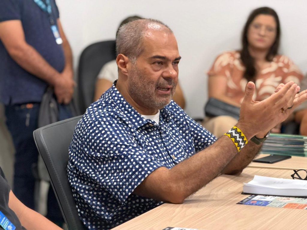 Um homem de camisa azul com estampa de poá fala durante uma reunião, gesticulando com as mãos. Ele usa uma pulseira artesanal com padrão geométrico em amarelo, preto e branco.
