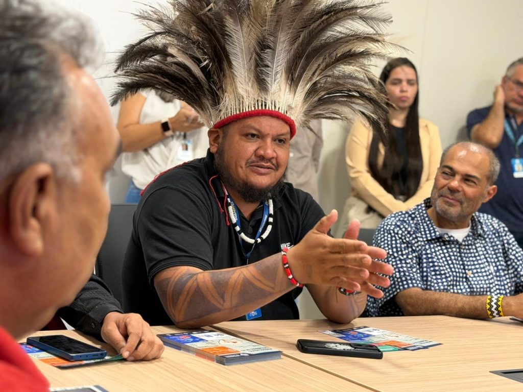 Um homem indígena de camiseta preta e cocar de penas discursa em uma mesa de reunião. Ele possui tatuagens de grafismos nos braços e usa colares e pulseiras artesanais. Ao lado, o homem da imagem anterior o observa atentamente.
