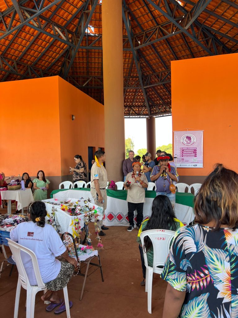 Fotografia em plano médio de uma cerimônia em um espaço coberto com teto de madeira e paredes laranjas. Ao centro, um casal de idosos indígenas fala ao microfone; eles usam colares e adornos tradicionais. À esquerda, um homem em uniforme bege observa. Em primeiro plano, pessoas sentadas de costas assistem à cena. Há uma mesa com toalha branca e detalhes geométricos em vermelho, além de um banner rosa à direita escrito "Para Mulheres".