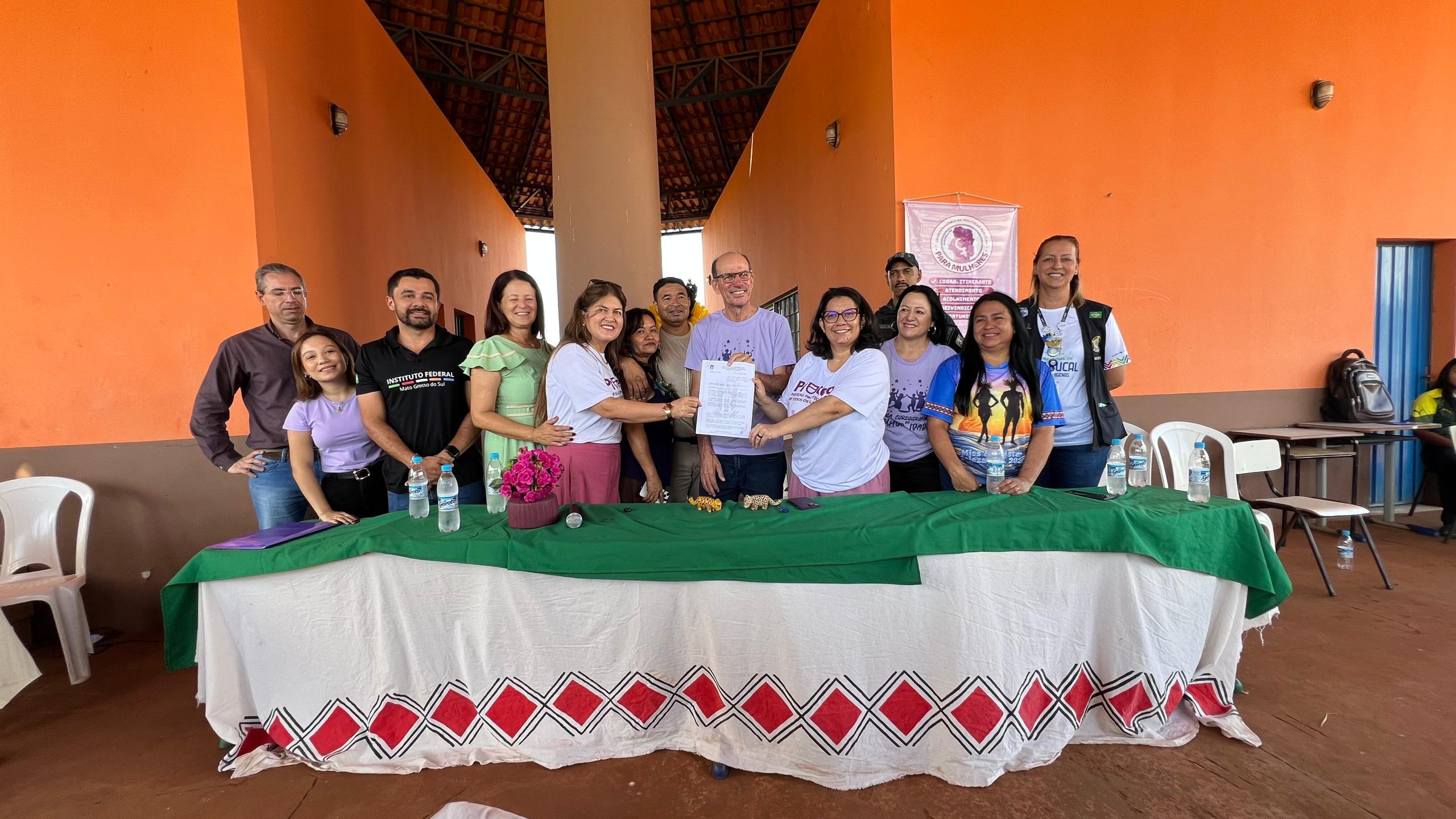 Fotografia de um grupo de cerca de 15 pessoas posadas para uma foto oficial atrás de uma mesa comprida. A mesa está coberta com uma toalha branca decorada com losangos vermelhos e um detalhe verde no topo. Duas mulheres ao centro seguram juntas um documento oficial branco. O grupo é diversificado, composto por homens e mulheres, incluindo representantes de forças de segurança e saúde. Ao fundo, a estrutura laranjada e o banner rosa da campanha "Para Mulheres".