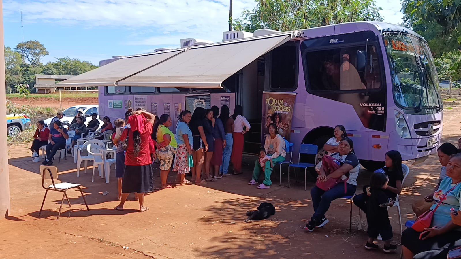 Fotografia externa de um ônibus lilás estacionado em um terreno de terra batida sob céu azul. O ônibus tem um toldo lateral estendido, criando uma área de sombra onde várias mulheres indígenas aguardam em fila ou sentadas em cadeiras plásticas brancas e azuis. Na lateral do ônibus, há adesivos de órgãos governamentais e o slogan "Todas por Todas". Ao fundo, árvores e vegetação rasteira.