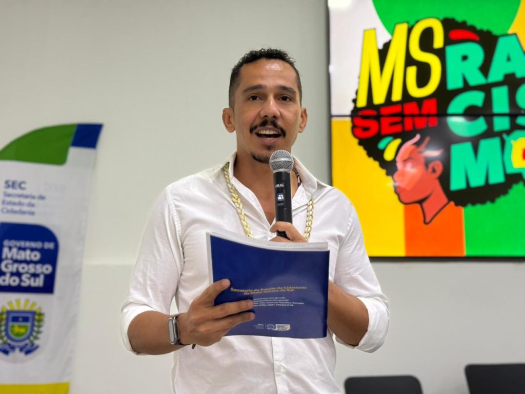 Homem jovem de pele parda, com barba e bigode curtos, fala ao microfone. Ele veste uma camisa social branca com os primeiros botões abertos, revelando uma corrente dourada grossa no pescoço. Em sua mão esquerda, segura um microfone preto; na direita, uma pasta azul com o logotipo da Secretaria de Estado da Cidadania de Mato Grosso do Sul. Ao fundo, à direita, há uma tela colorida com a ilustração de uma mulher negra de perfil e a frase "MS RACISMO NÃO". À esquerda, aparece parte de um banner do Governo do Estado de Mato Grosso do Sul.