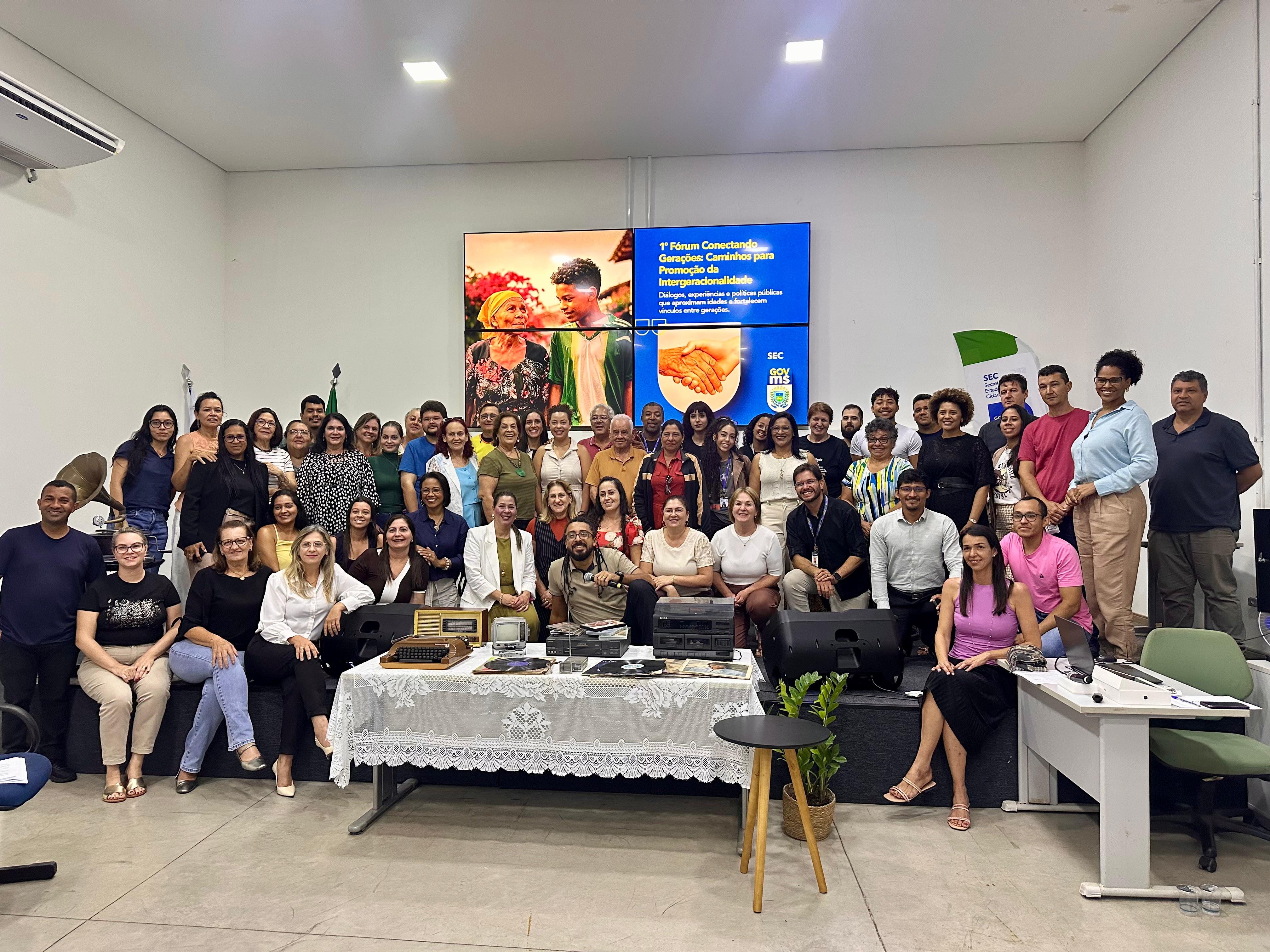 Fotografia oficial de encerramento com um grande grupo de participantes reunidos no palco e à frente dele. São dezenas de pessoas de diversas idades, homens e mulheres, sorrindo para a câmera. Ao centro, no chão, estão alguns objetos antigos que compunham a cenografia. O telão ao fundo permanece ligado com a logomarca do evento, e a iluminação do auditório é clara e uniforme.