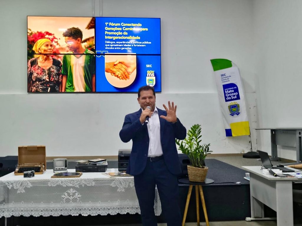 Um homem de terno azul escuro e camisa branca fala ao microfone no centro do palco. Ele faz um gesto com a mão esquerda aberta. Ao fundo, o painel digital azul destaca o tema da intergeracionalidade. No palco, à esquerda, há uma mesa com objetos antigos e, à direita, um banner da Secretaria de Estado de Cidadania (SEC) e do Governo de Mato Grosso do Sul.