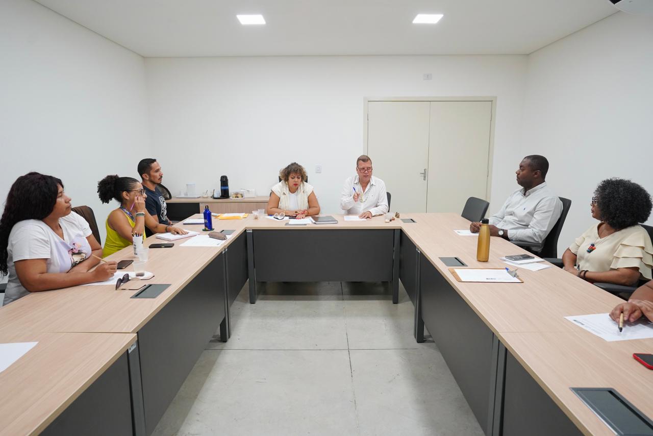 Sete pessoas estão sentadas ao redor de uma grande mesa de madeira em formato de "U" em uma sala de reuniões clara. Ao centro, no topo do "U", uma mulher e um homem branco parecem conduzir a conversa, com papéis e canetas à frente. Nas laterais da mesa, quatro mulheres negras e um homem negro participam da reunião, alguns tomando notas e outros observando atentamente. O ambiente é profissional, com paredes brancas, iluminação embutida no teto e uma porta fechada ao fundo. Sobre a mesa, veem-se garrafas de água, celulares e pastas de documentos.