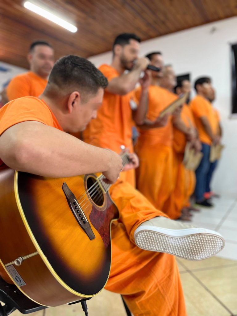 Close-up lateral de um homem de uniforme laranja tocando violão. Ao fundo, outros internos em fila aparecem cantando, com foco voltado para o instrumento em primeiro plano.