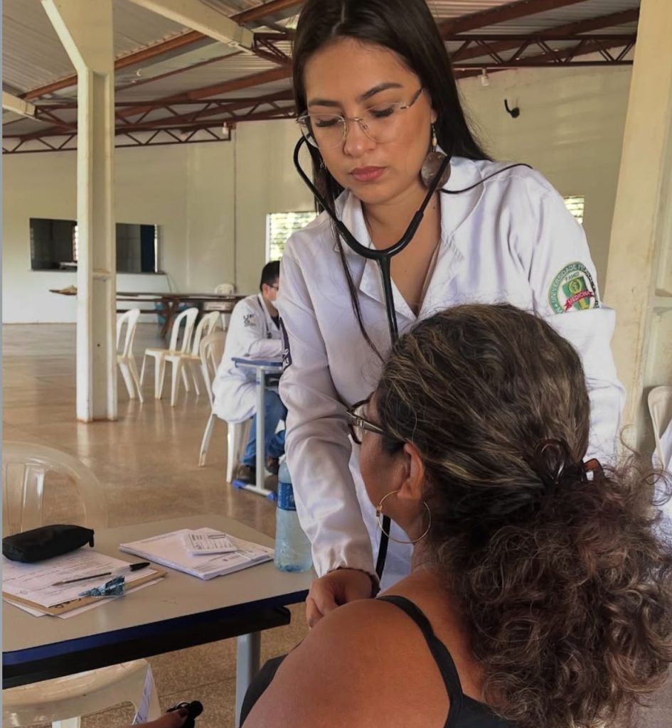 Uma profissional de saúde, vestindo jaleco branco e óculos, utiliza um estetoscópio para realizar um atendimento médico. Ela está inclinada sobre uma paciente de costas, que possui cabelos cacheados e usa óculos. O atendimento ocorre em um ambiente amplo e arejado.