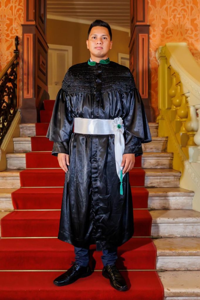 Um homem jovem posa para uma foto formal em uma escadaria de mármore com tapete vermelho. Ele veste uma beca de formatura preta com uma faixa branca na cintura e detalhes em verde na gola. O ambiente ao fundo possui paredes decoradas em tons de laranja e dourado.