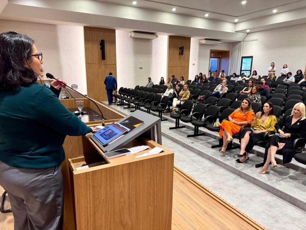 Vista lateral do auditório a partir do palco. A palestrante de blusa verde aparece em primeiro plano, de costas, enquanto a plateia, composta majoritariamente por mulheres, acompanha a apresentação sentada em poltronas pretas.