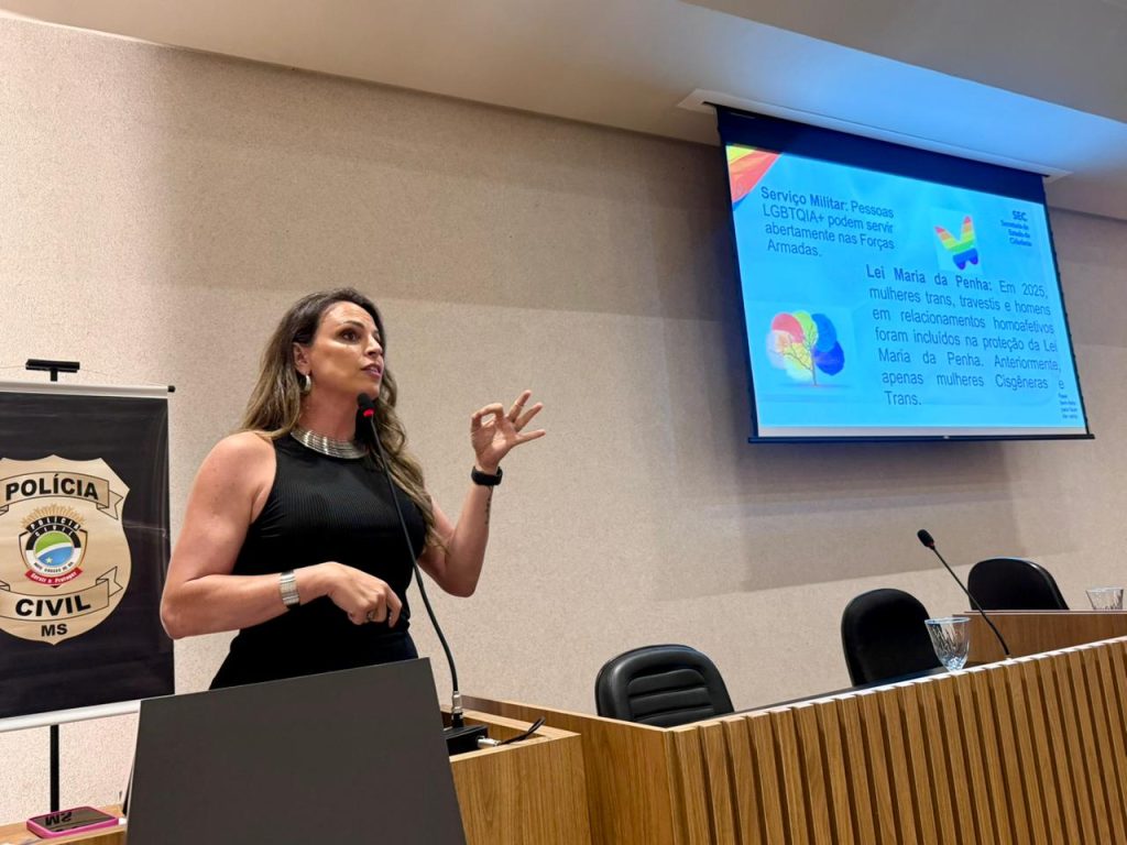 Uma mulher de cabelos longos e blusa preta sem mangas fala ao microfone no púlpito. Ao fundo, um telão exibe informações sobre a "Lei Maria da Penha" e atualizações sobre a proteção de mulheres trans e homens trans.