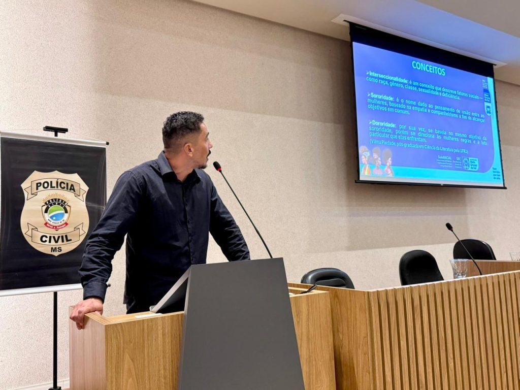Um homem de camisa preta fala ao púlpito, olhando para o lado. No telão ao fundo, lê-se o título "CONCEITOS" seguido de definições sobre Interseccionalidade, Sororidade e Dororidade.