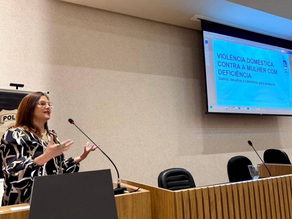 Uma mulher de óculos e blusa estampada em preto e branco gesticula com as mãos enquanto fala. O telão ao fundo exibe o título "VIOLÊNCIA DOMÉSTICA CONTRA A MULHER COM DEFICIÊNCIA".