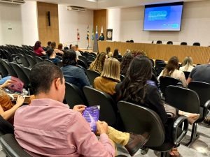 Visão geral do auditório de trás para frente. O homem de camisa rosa aparece em primeiro plano, e o foco se estende até o palco, onde a palestrante conduz o evento diante de uma plateia atenta.