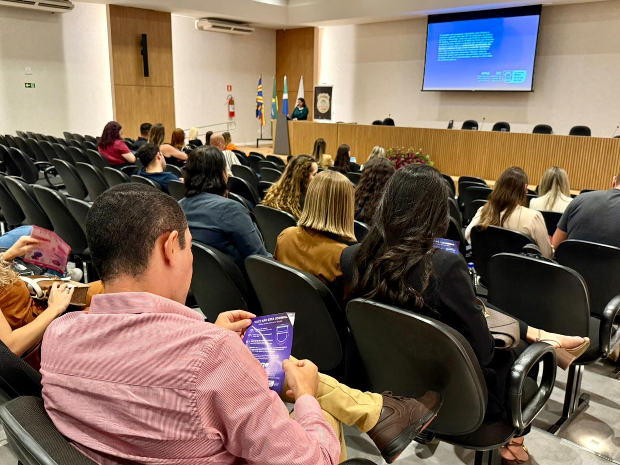 Visão geral do auditório de trás para frente. O homem de camisa rosa aparece em primeiro plano, e o foco se estende até o palco, onde a palestrante conduz o evento diante de uma plateia atenta.