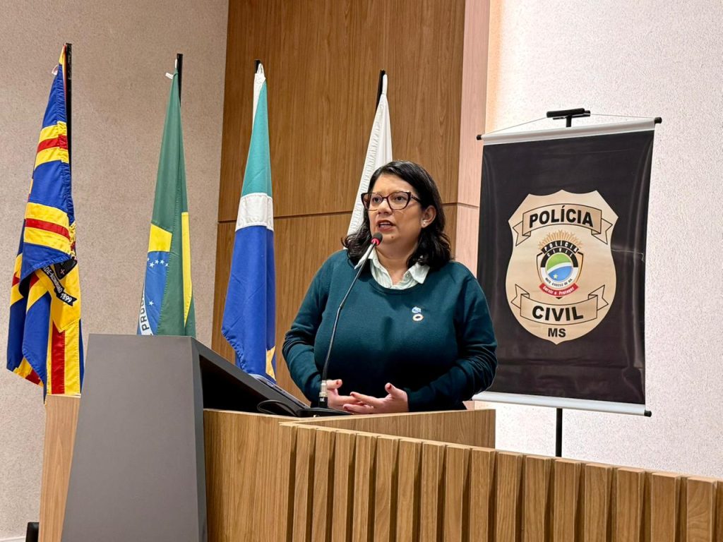 Uma mulher de óculos e blusa verde escura discursa em um púlpito de madeira. Ao fundo, as bandeiras do Brasil e do Mato Grosso do Sul, além de um banner com o brasão da Polícia Civil de MS.