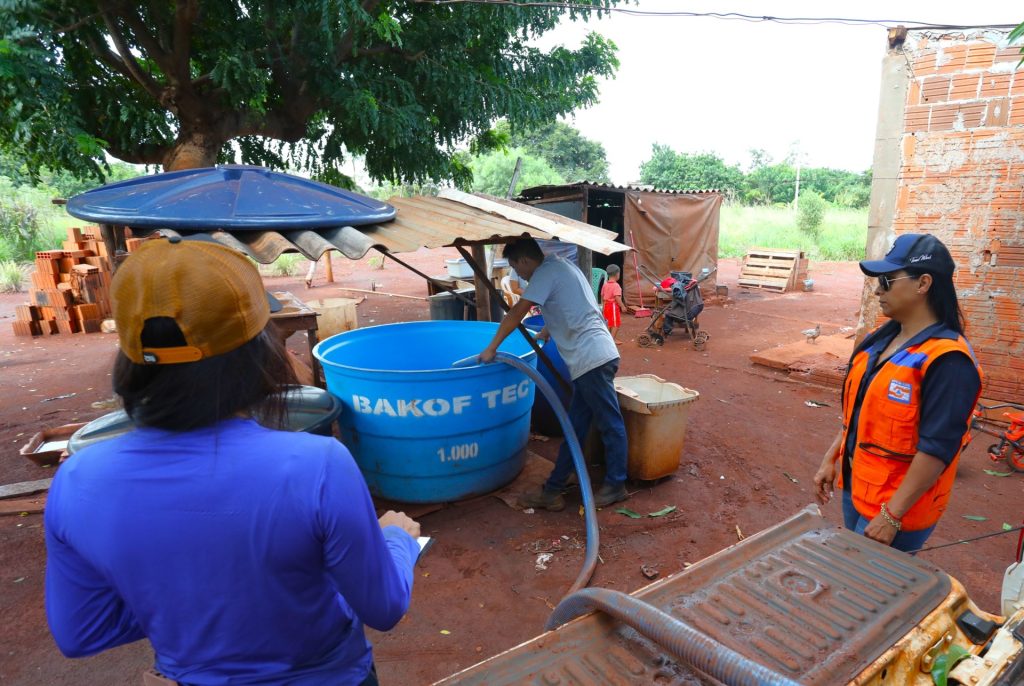 Vista aérea parcial de um quintal de terra vermelha onde um homem direciona uma mangueira para encher uma grande caixa d'água azul com a marca "BAKOF TEC". Duas mulheres, uma vestindo blusa azul e boné amarelo (de costas) e outra com colete laranja da Defesa Civil (de perfil), acompanham a ação.