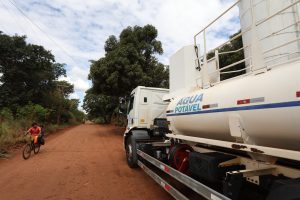 Em primeiro plano, a lateral de um caminhão-pipa branco parado em uma estrada de terra vermelha. No tanque do veículo, lê-se a inscrição "ÁGUA POTÁVEL" em azul. À esquerda, na estrada arborizada, um homem anda de bicicleta sob um céu com nuvens brancas.