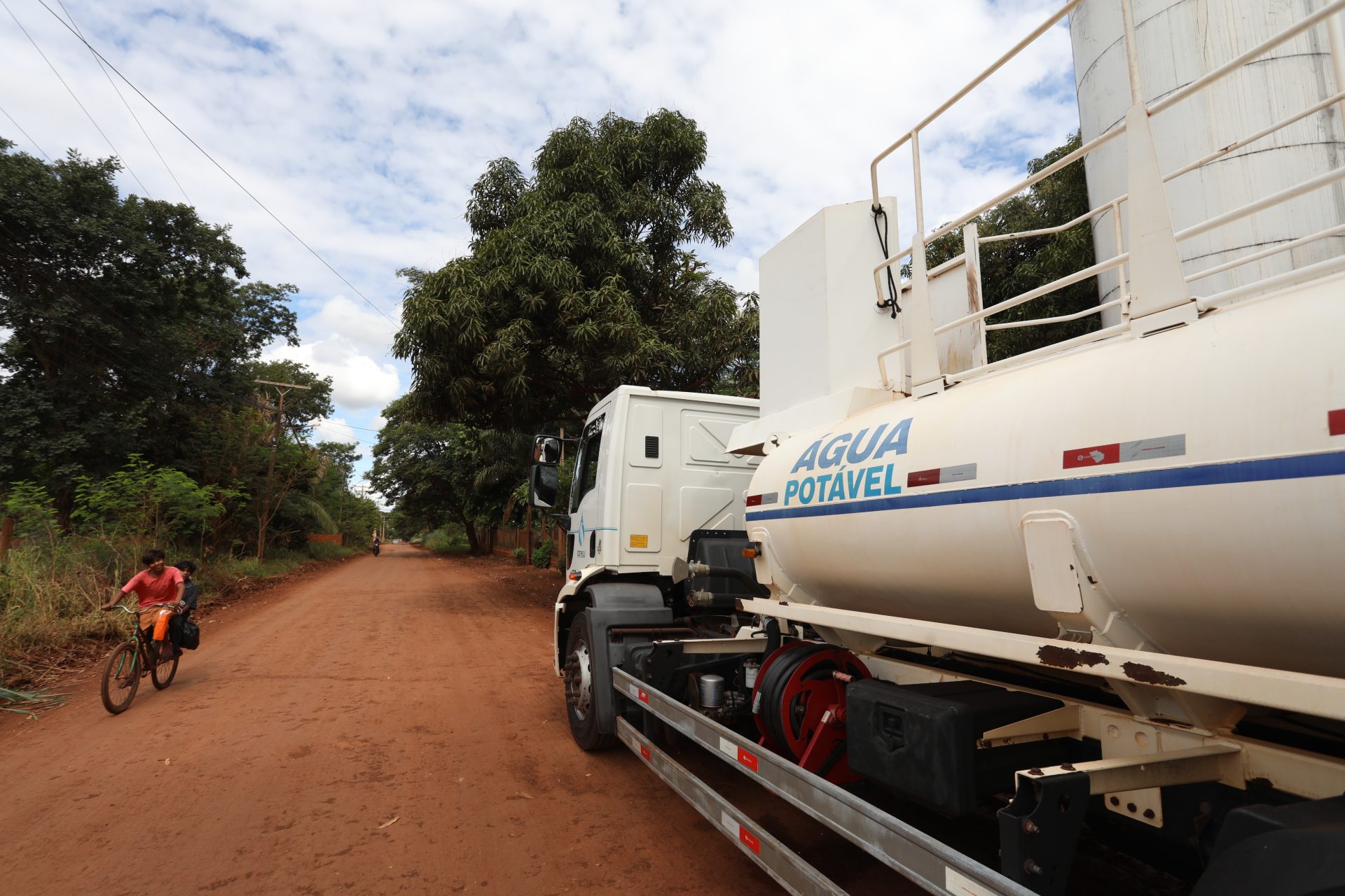 Em primeiro plano, a lateral de um caminhão-pipa branco parado em uma estrada de terra vermelha. No tanque do veículo, lê-se a inscrição "ÁGUA POTÁVEL" em azul. À esquerda, na estrada arborizada, um homem anda de bicicleta sob um céu com nuvens brancas.