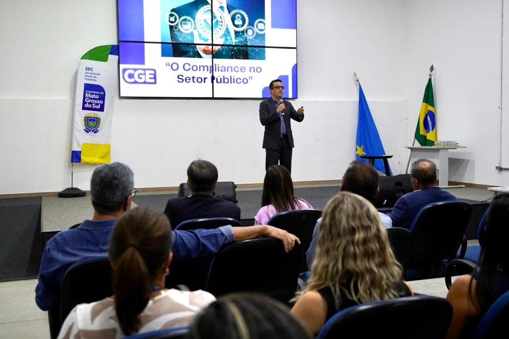 Um homem de terno e óculos faz uma apresentação em um palco. Atrás dele, um telão exibe a frase "O Compliance no Setor Público" ao lado do logotipo da CGE. No palco, há também um banner da SEC e as bandeiras de Mato Grosso do Sul e do Brasil. Em primeiro plano, vemos as cabeças e ombros de algumas pessoas da plateia de costas.