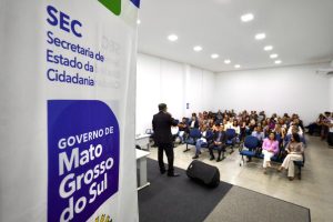 Foto tirada de trás de um banner da Secretaria de Estado da Cidadania (SEC) e do Governo de Mato Grosso do Sul, que ocupa o lado esquerdo da imagem. Ao fundo, um homem de terno fala para uma plateia atenta sentada em poltronas azuis em um auditório branco e bem iluminado.