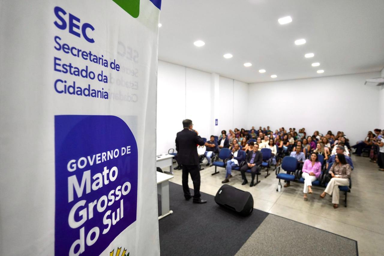 Foto tirada de trás de um banner da Secretaria de Estado da Cidadania (SEC) e do Governo de Mato Grosso do Sul, que ocupa o lado esquerdo da imagem. Ao fundo, um homem de terno fala para uma plateia atenta sentada em poltronas azuis em um auditório branco e bem iluminado.