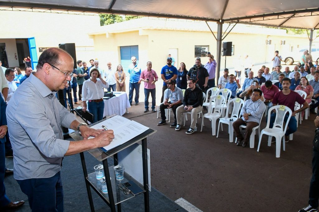 Sob uma tenda de eventos, um homem calvo e de óculos, vestindo camisa cinza, assina um documento oficial posicionado em um púlpito. Ao fundo, uma plateia mista sentada em cadeiras brancas de plástico e outras pessoas em pé assistem solenemente ao ato.