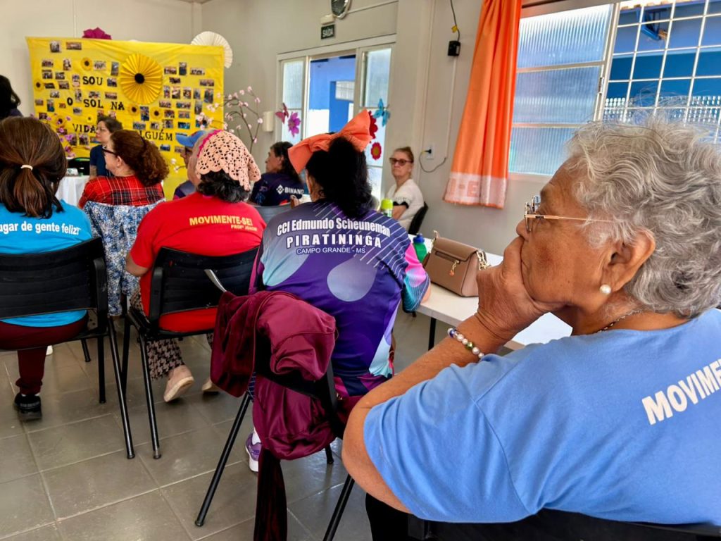 Foto em ângulo diagonal mostrando idosos sentados em cadeiras assistindo a uma atividade. Em primeiro plano, à direita, uma mulher idosa de cabelos grisalhos e blusa azul apoia a mão no queixo enquanto observa atentamente. À sua frente, outras pessoas estão de costas; uma delas veste uma camiseta roxa e azul escrito "CCI Edmundo Scheunemann - PIRATININGA - Campo Grande - MS". Ao fundo, destaca-se um painel decorativo amarelo com fotos e flores artesanais.
