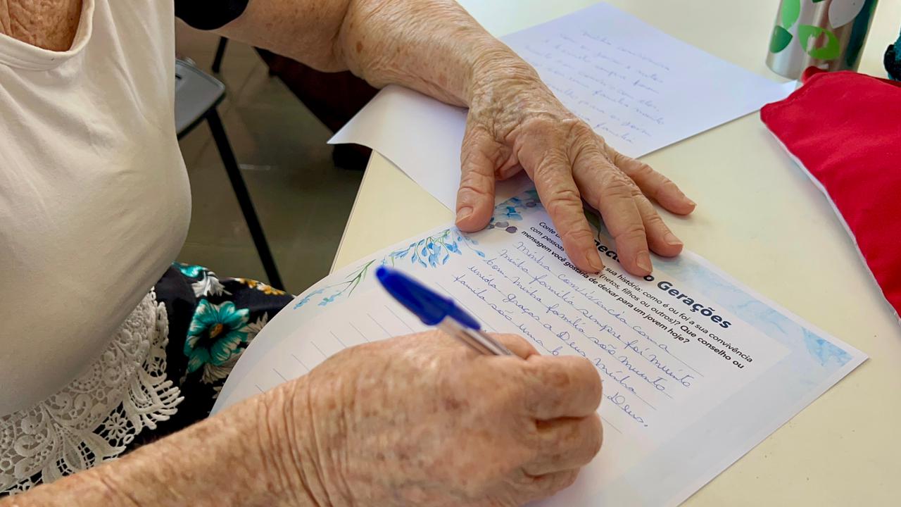 Imagem em plano detalhado e focado nas mãos de uma pessoa idosa escrevendo com uma caneta azul em uma folha de papel sobre a mesa. O papel timbrado possui detalhes florais azuis e traz o título "Conectando Gerações", seguido por uma mensagem manuscrita com letra cursiva que fala sobre convivência familiar e gratidão a Deus.