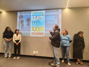 Cinco pessoas estão alinhadas à frente de uma parede cinza onde é projetado um slide com o título "Brincadeiras Africanas Divertidas e Educativas". Um homem ao centro, vestindo jaqueta preta, aponta para a projeção enquanto fala, observado por duas mulheres à esquerda e duas à direita.