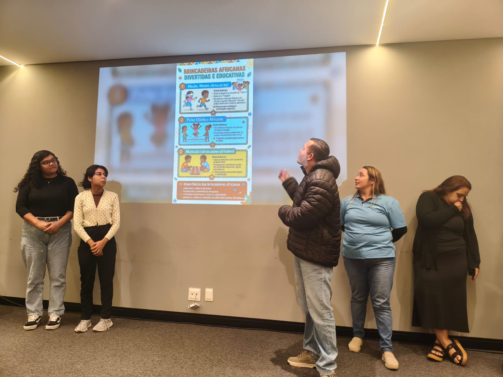 Cinco pessoas estão alinhadas à frente de uma parede cinza onde é projetado um slide com o título "Brincadeiras Africanas Divertidas e Educativas". Um homem ao centro, vestindo jaqueta preta, aponta para a projeção enquanto fala, observado por duas mulheres à esquerda e duas à direita.
