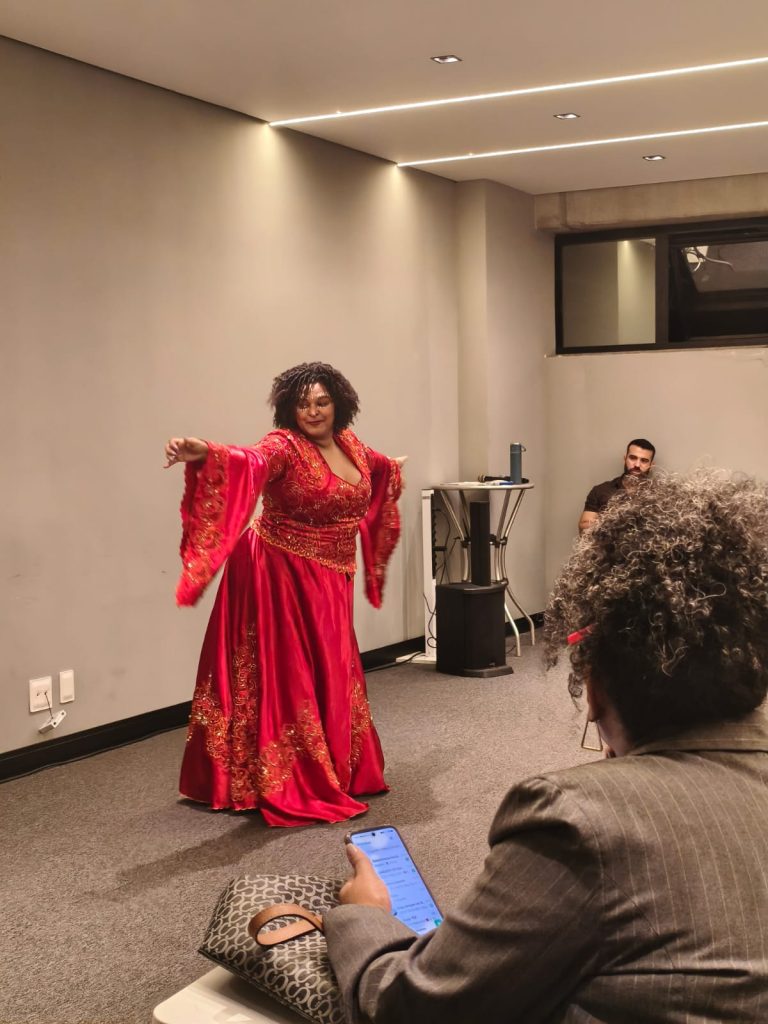 Uma mulher negra com cabelos cacheados dança expressivamente no centro de uma sala, vestindo um imponente vestido longo vermelho com detalhes e bordados dourados. A cena é registrada em primeiro plano pelas costas de uma pessoa na plateia que segura um celular, enquanto ao fundo um homem observa a apresentação sentado.