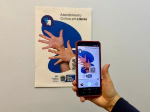 Close de uma mão segurando um smartphone que aponta para um cartaz na parede branca. O cartaz diz "Atendimento Online em Libras" e exibe um QR Code e o logotipo do Governo de Mato Grosso do Sul.