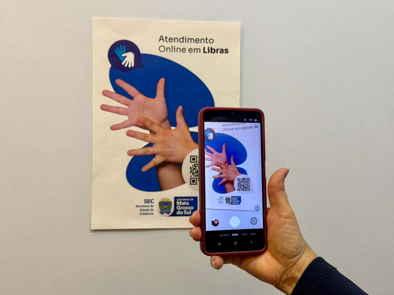 Close de uma mão segurando um smartphone que aponta para um cartaz na parede branca. O cartaz diz "Atendimento Online em Libras" e exibe um QR Code e o logotipo do Governo de Mato Grosso do Sul.