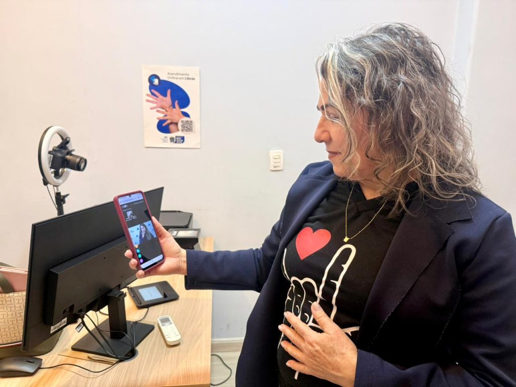 Uma mulher com camiseta preta (estampada com o sinal de "eu te amo" em Libras) segura um celular mostrando uma videochamada. Ela está ao lado de uma mesa com monitor e um ring light acoplado a uma câmera.